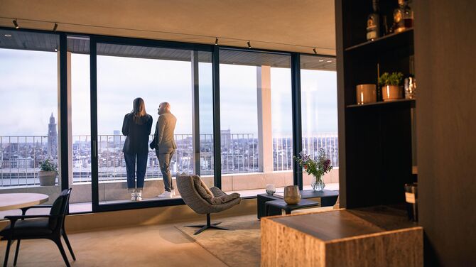 Alt : Vue intérieure de l’appartement moderne avec de larges portes coulissantes et deux personnes sur un balcon.
